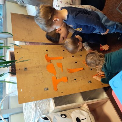 Trójka dzieci przyczepia papierowe narzędzia do drewnianej płyty. Kliknięcie w obrazek spowoduje powiększenie.