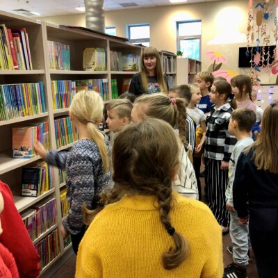 Uczniowie stoją przy regałach i słuchają Pani Bibliotekarki, która opowiada im o układzie książek na półkach. Kliknięcie w obrazek spowoduje powiększenie.