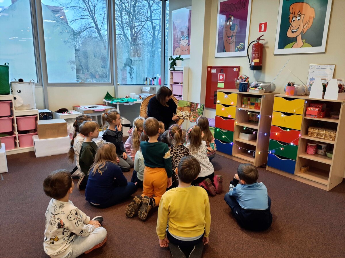 Przedszkolaki w skupieniu słuchają książki czytanej przez Panią Bibliotekarkę.