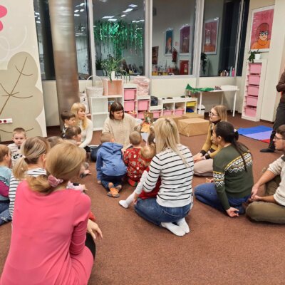Dzieci wraz z opiekunami siedzą na dywanie i w skupieniu słuchają książki czytanej przez Panie Bibliotekarki. Kliknięcie w obrazek spowoduje powiększenie.