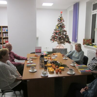 grupa uśmiechniętych osób dookoła stołu z logo programu DKK, stroikiem, pomarańczami, słodyczami i herbatą; w tle udekorowana i podświetlona świąteczna choinka i regały z książkami. Kliknięcie w obrazek spowoduje powiększenie.