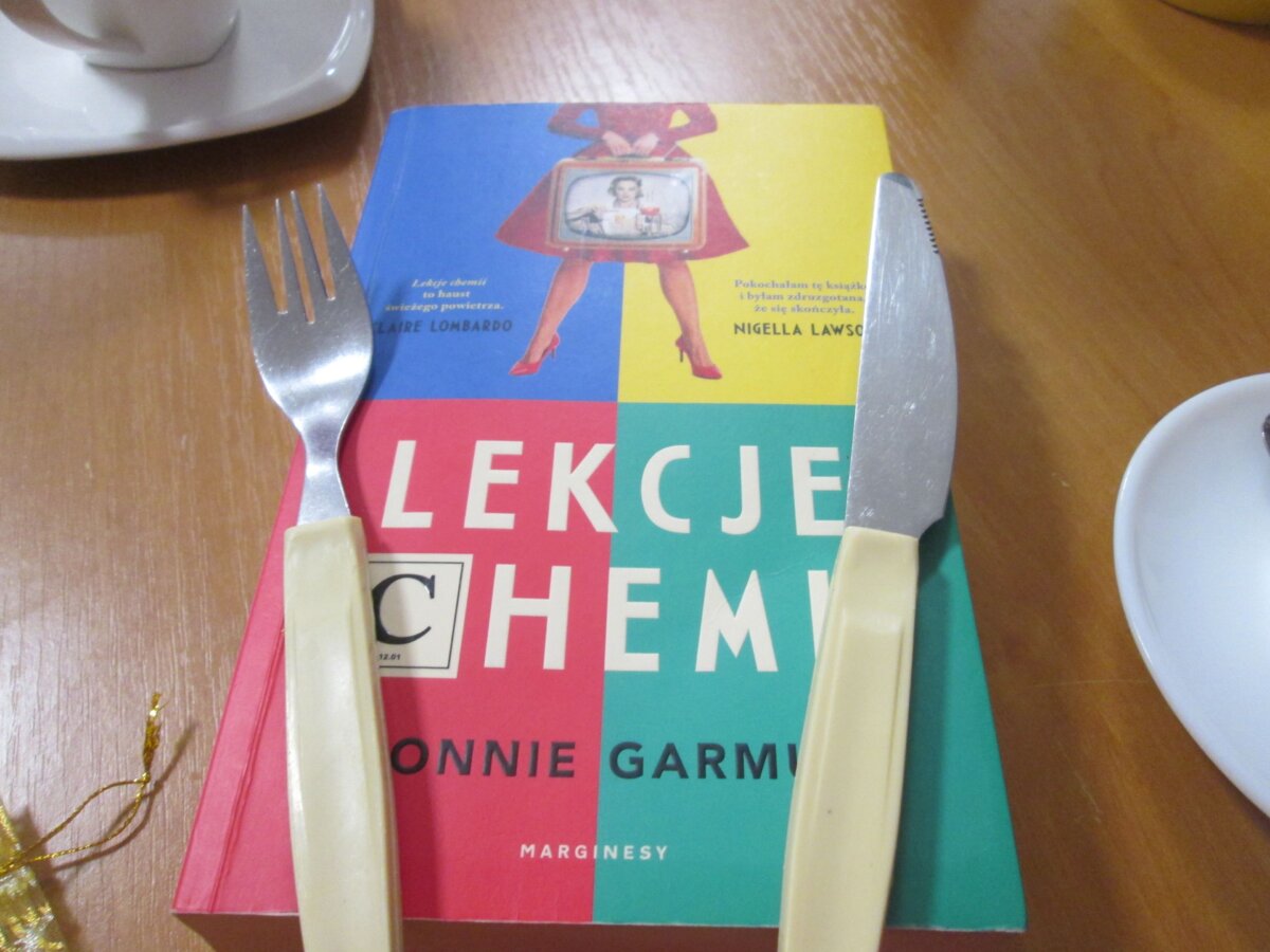 zbliżenie na okładkę książki Bonnie Garmus pt. „Lekcje chemii” z nożem i widelcem na jej wierzchu po lewej i prawej stronie brzegów okładki; obok fragmenty filiżanek.