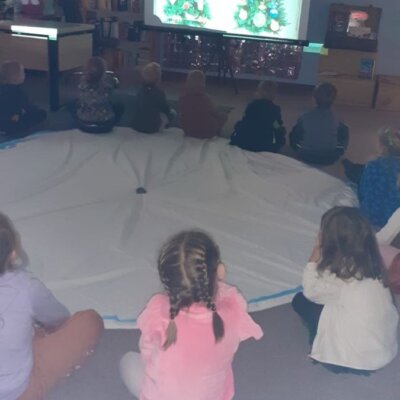 Dzieci siedzą na dywanie i oglądają prezentację o choince, przygotowaną przez bibliotekarką. Kliknięcie w obrazek spowoduje powiększenie.