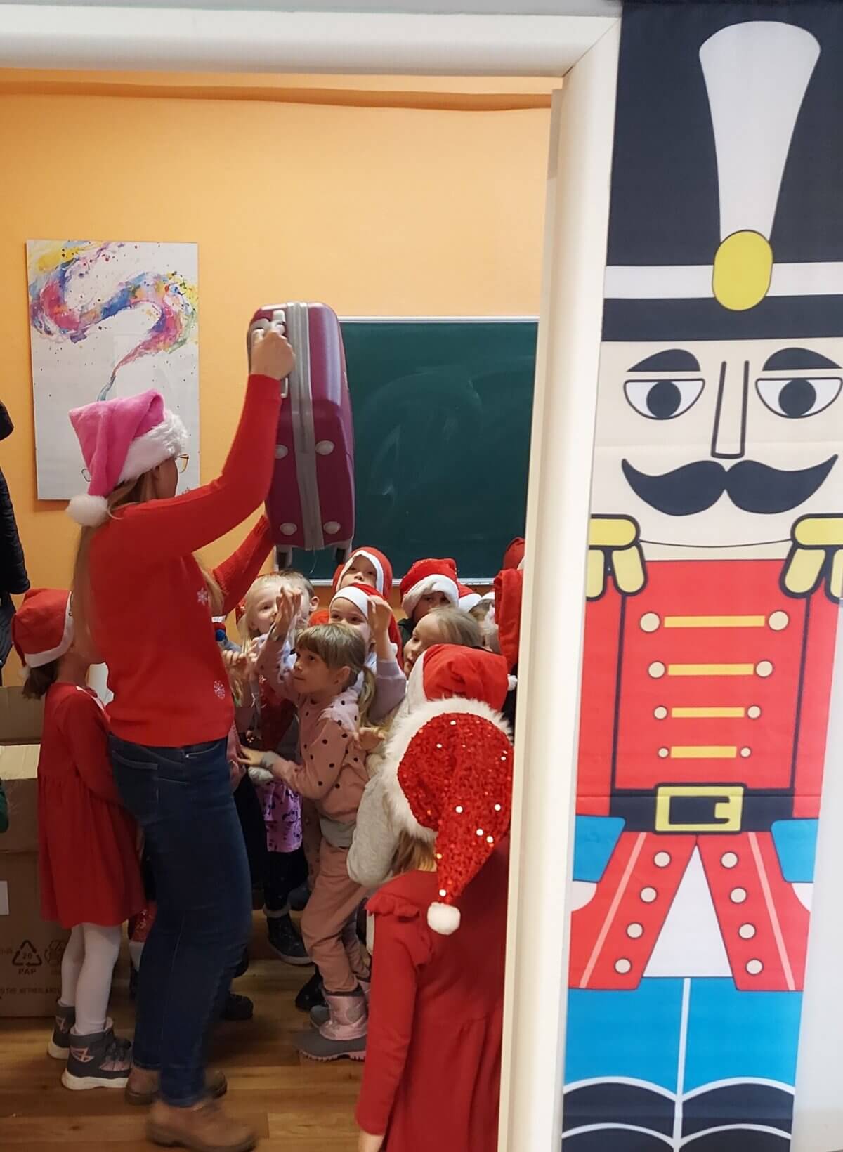 Z prawej strony zdjęcia świąteczny żołnierz w barwach czerwonych, niebieskich i żółtych. Lewa część zdjęcia przedstawia grupkę dzieci w czerwonych mikołajowych czapkach patrzących na osobę dorosłą trzymającą w górze różową walizkę.