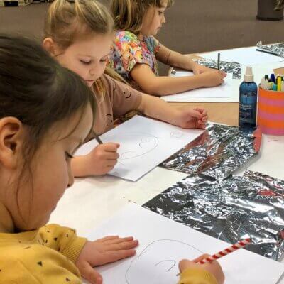 Trzy dziewczynki siedzą przy stole i na kartkach papieru rysują różne emocje. Kliknięcie w obrazek spowoduje powiększenie.