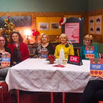 Siedem kobiet siedzi przy stole. Są radosne i uśmiechnięte. Eksponują omawianą książkę. W tle po lewej stronie udekorowana choinka. Kliknięcie w obrazek spowoduje powiększenie.