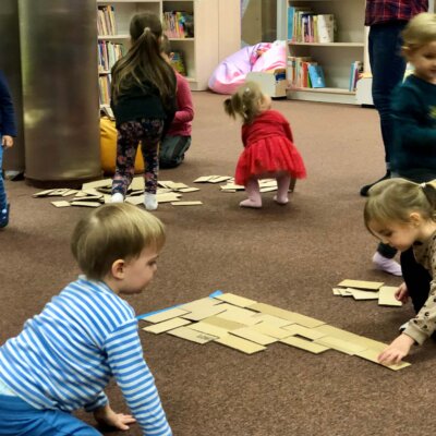 Grupa dzieci układa kartonowe cegiełki na ziemi, tworząc w ten sposób front domku. Kliknięcie w obrazek spowoduje powiększenie.
