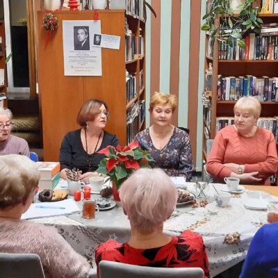 Grupa dziewięciu osób skupiona wokół stołu siedzi zasłuchana w opowieść kobiety zwracającej się do słuchaczy. Na stole widoczne ułożone książki – biografie oraz zbiory utworów Konstantego Ildefonsa Gałczyńskiego. Kliknięcie w obrazek spowoduje powiększenie.