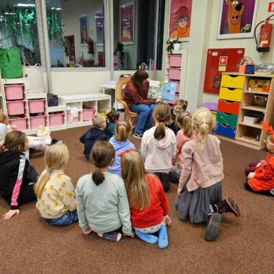 Dzieci siedzą na dywanie i w skupieniu słuchają książki czytanej przez Panią Bibliotekarkę. Kliknięcie w obrazek spowoduje powiększenie.