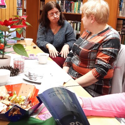Zdjęcie przedstawia ożywioną dyskusję pomiędzy dwoma kobietami. Siedząca u szczytu stołu członkini DKK rozmawia z sąsiadką wyciągając przed siebie dłonie. Na pierwszym planie otwarta książka trzymana przez osobę niewidoczną na zdjęciu.