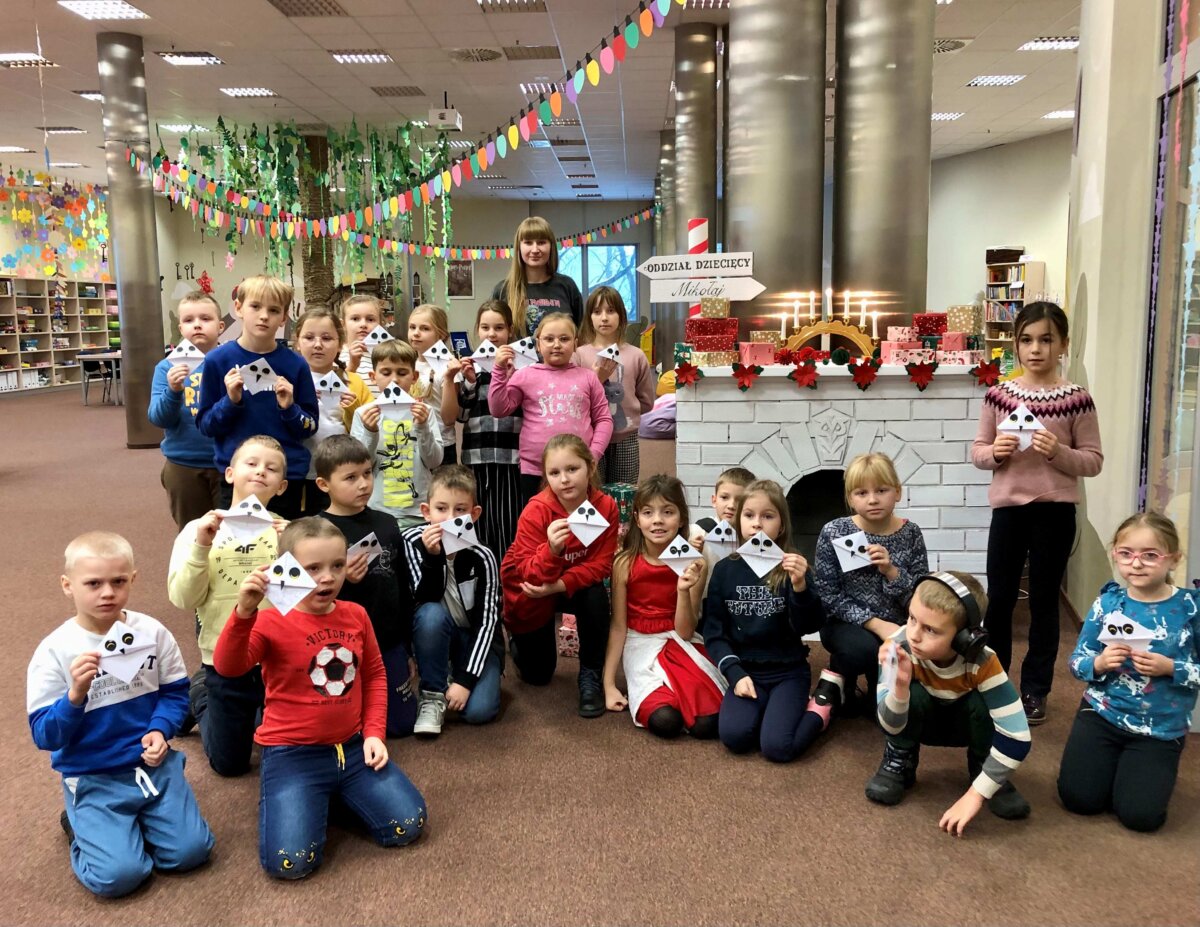Dzieci wraz z Panią bibliotekarką pozują do wspólnego zdjęcia na tle kominka i prezentów. Każde z dzieci trzyma w dłoniach wykonaną zakładkę do książki.