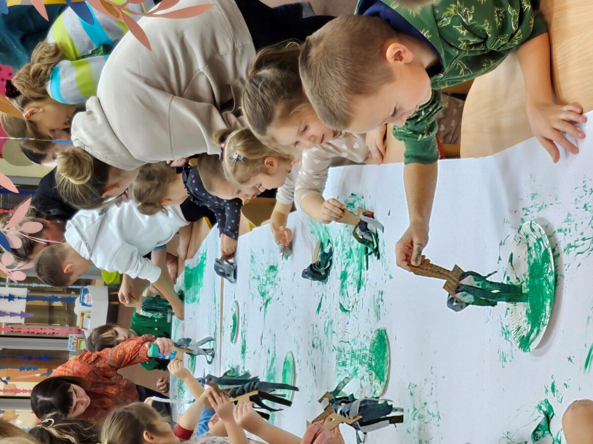 Używając własnoręcznie wykonanych pędzli, dzieci zamalowują biały arkusz kartki zieloną farbą.