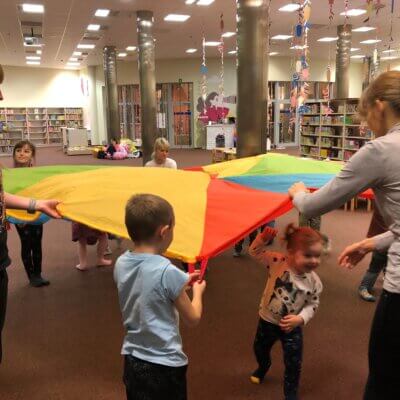 Pani Bibliotekarka wraz z dziećmi trzyma chustę animacyjną, pod którą chowają się najmłodsi. Kliknięcie w obrazek powoduje powiększenie.