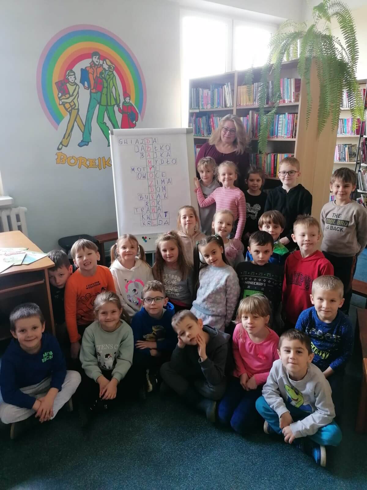 Grupa dzieci z osobą dorosłą na tle ściany z logo biblioteki „Borejkowo”, i regałów z książkami. Przy tablicy z rozwiązaną krzyżówką z hasłem „Dbajmy o zdrowie”.