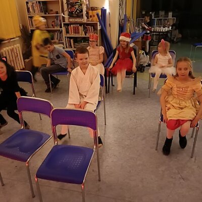 9 dzieci w strojach karnawałowych grających w grę pod tytułem „Kareta”, 7 dzieci siedzi na niebieskich krzesłach, dwa dodatkowe krzesła są puste, a dwójka dzieci biega wokół pozostałych. Kliknięcie powoduje powiększenie zdjęcia.