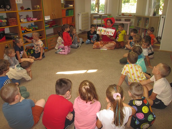 Salka przedszkola. 22 dzieci na dywanie, a wśród nich dorosła ubrana w czerwone akcenty z maską w kształcie jabłka, czytająca bajkę pt. „Bardzo głodna gąsienica".