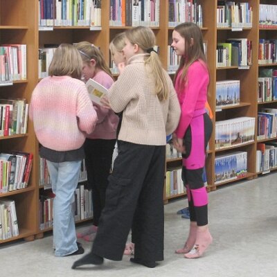 6 dziewczynek przeglądających książkę przy ścianie z regałami pełnymi książek. Kliknięcie powoduje powiększenie zdjęcia.