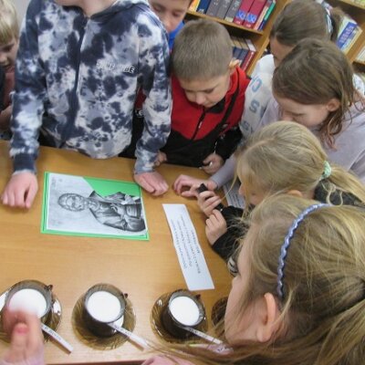 Grupa dzieci przyglądająca się kartce z tekstem, zdjęciu świętego Andrzeja i trzy filiżanki brązowe filiżanki z sola na biurku. Kliknięcie powoduje powiększenie zdjęcia.