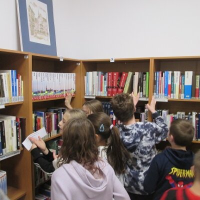 Grupa dzieci w kąciku z regałami pełnymi książek. Na regale duża antyrama z fotografia, a na brzegach regałów rzymskie cyfry. Kliknięcie powoduje powiększenie zdjęcia.