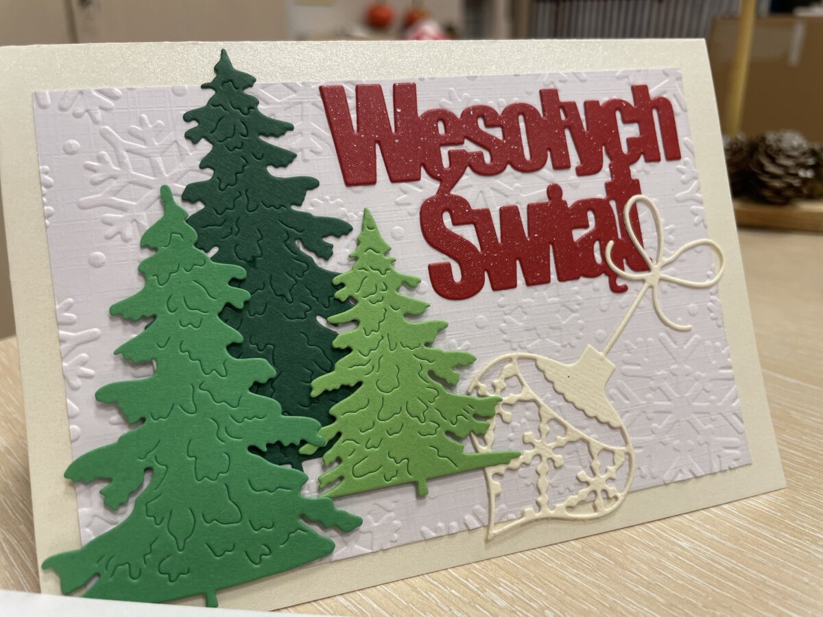 Świąteczna kartka 3D z czerwonym napisem Wesołych Świąt, trzy zielone choinki, każda w innym kolorze zieleni i bombka w kolorze ecru wytłoczone z papieru, przyklejone na białym wytłoczonym w gwiazdki śniegu tle.