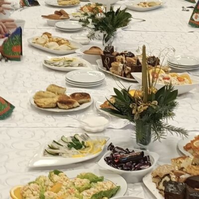 Stół nakryty białym obrusem, na nim półmiski z potrawami wigilijnymi i dwa stroiki świąteczne z czerwonymi świeczkami. Białe nakrycia stołu. Kliknięcie powoduje powiększenie zdjęcia.