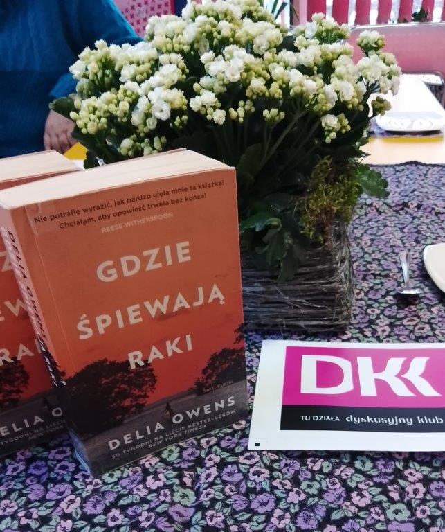 Tytułowe. Na stole książka obok koszyka z kwiatami i napis DKK.