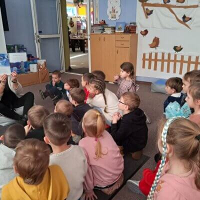 Dzieci siedzą na dywanie, bibliotekarka czyta dzieciom książkę „Kraina Lodu” i pokazuje ilustracje.