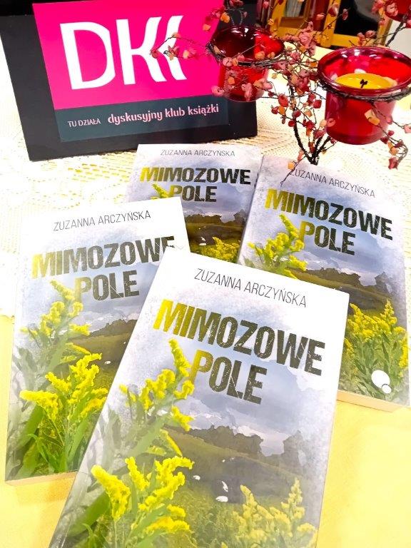 Na stole leżą cztery książki „Mimozowe pole” Zuzanny Arczyńskiej Nad nimi stoi tabliczka z logo DKK – dyskusyjnego klubu książki i dekoracyjny świecznik