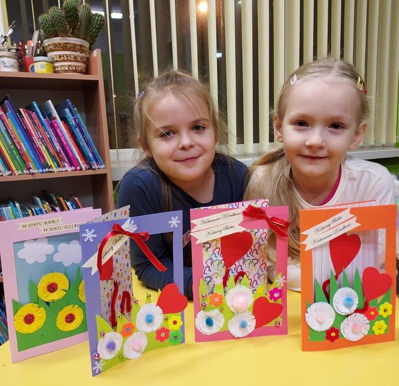 Dwójka dzieci na tle kartek wykonanych z kolorowego papieru z okazji Dnia Babci i Dnia Dziadka, ozdobionych kwiatkami, serduszkami, koralikami i pomponikami.