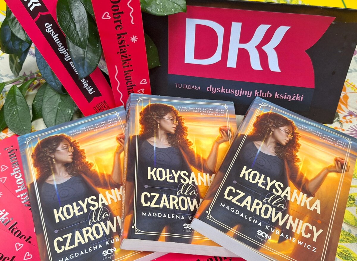 Na stole leżą trzy książki Magdaleny Kubasiewicz „Kołysanka dla czarownicy”. Nad nimi stoi tabliczka z logo DKK – dyskusyjnego klubu książki.