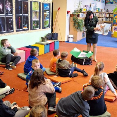 W bibliotece na dywanie siedzą dzieci i patrzą na dianę, która ma na sobie galowy mundur myśliwski i trzyma zdjęcie lisa. W tle widać regały z książkami. Kliknięcie w obrazek spowoduje powiększenie.