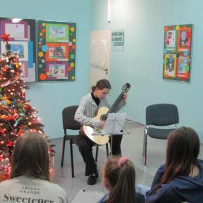 Dziewczyna w pasiastej koszuli gra na gitarze akustycznej z pochyloną głową, dookoła niej zasłuchana publiczność. Kliknięcie w obrazek powoduje powiększenie.