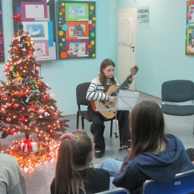 Dziewczynka trzymająca gitarę klasyczną bardzo uważnie śledzi nuty na pulpicie, obok niej stoi rozświetlona bożonarodzeniowa choinka. Kliknięcie w obrazek powoduje powiększenie.