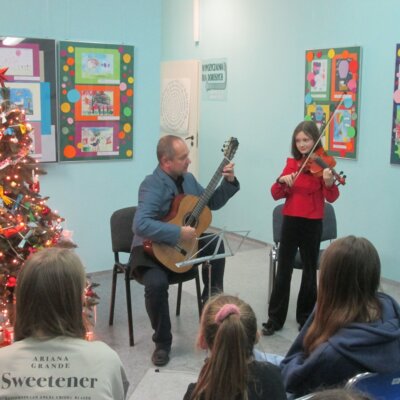 Duet muzyczny występuje w świątecznej scenerii: młoda skrzypaczka ubrana w czarne spodnie i błyszczącą czerwoną bluzkę patrzy z uśmiechem na swego nauczyciela, który gra na gitarze klasycznej. Kliknięcie w obrazek powoduje powiększenie.