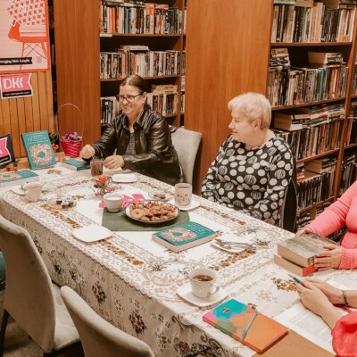 W bibliotece ma miejsce spotkanie Dyskusyjnego Klubu Książki. Na stole leżą filiżanki i ciastka. Jedna z kobiet opowiada, inne patrzą na nią. Niektóre się uśmiechają. Kliknięcie powoduje powiększenie zdjęcia.