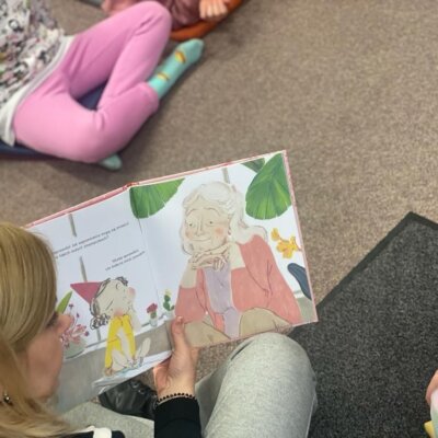 Zdjęcie przedstawia prowadzącą, która czyta dzieciom książkę i pokazuje ilustracje. Kliknięcie powoduje powiększenie zdjęcia.