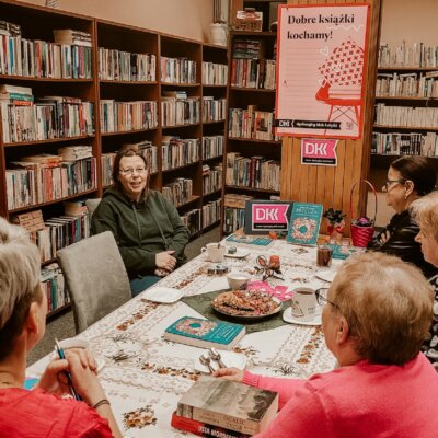 Wnętrze biblioteki. W tle regały z książkami. Trwa spotkanie DKK. Kobiety siedzą przy stole. Na pierwszym planie jedna z nich gestykuluje. Kliknięcie powoduje powiększenie zdjęcia.
