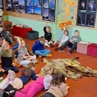 Dzieci siedzą na dywanie, kilkoro z nich podnosi ręce. Przed nimi leżą skóry zwierząt i stoi diana, która pokazuje im zdjęcia. Kliknięcie w obrazek spowoduje powiększenie.