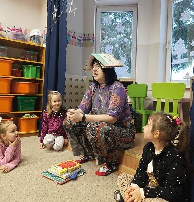 Bibliotekarka we wzorzystych ubraniach z książką na głowie, siedząca na podeście, otoczona dziećmi. Klikniecie powoduje powiększenie zdjęcia.