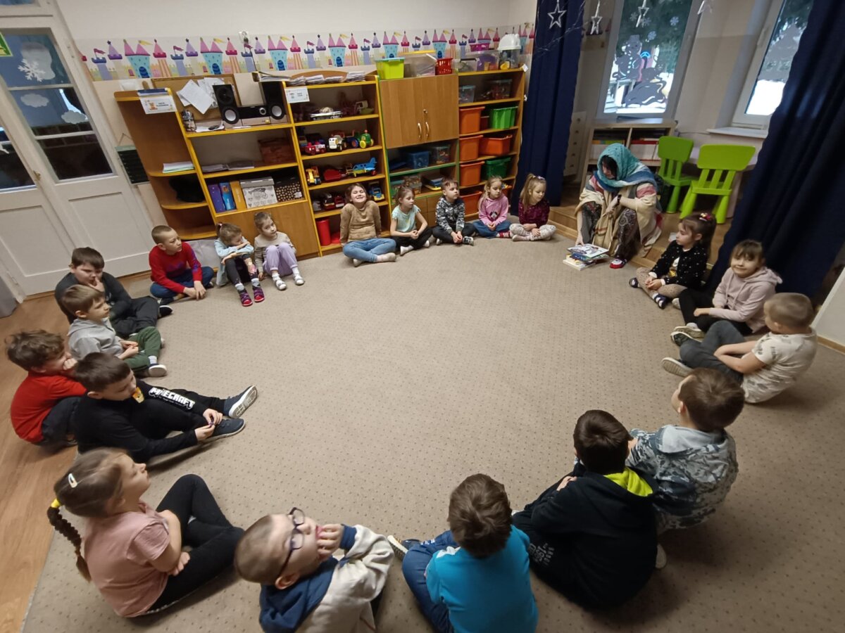 Grupa dzieci w w przedszkolu siedząca na kremowym dywanie razem z bibliotekarka przebraną za babcię.