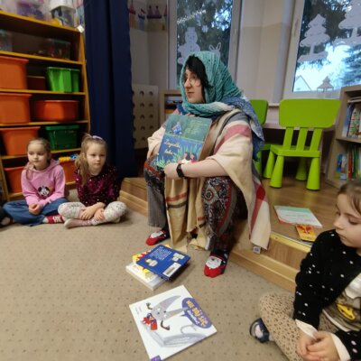Bibliotekarka siedząca na podeście przebrana za babcię w niebieskiej chuście i kremowym poncho, obok niej dzieci. Kliknięcie powoduje powiększenie zdjęcia.