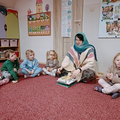 Bibliotekarka przebrana za babcię w niebieskiej chuście i kremowym poncho siedząca razem z piątką dzieci na czerwonym dywanie. Kliknięcie powoduje powiększenie zdjęcia.