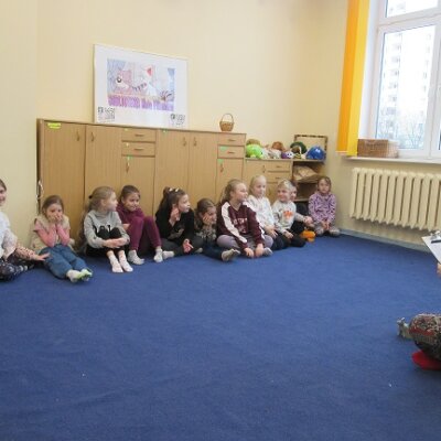 Jedenastka dzieci siedząca pod niską meblościanką na granatowym dywanie. Na przeciw dzieci czarnowłosa bibliotekarka siedząca po turecku. Klikniecie powoduje powiększenie zdjęcia.