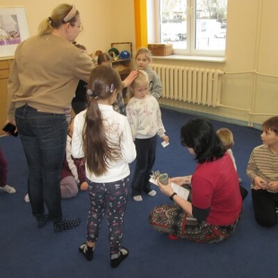 Grupa dzieci z nauczycielką stojące wokół bibliotekarki na granatowym dywanie. Kliknięcie powoduje powiększenie zdjęcia.