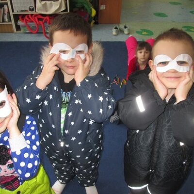 Trójka dzieci stojąca na granatowym dywanie przykładająca do twarzy skończone maski karnawałowe. Klikniecie powoduje powiększenie zdjęcia.