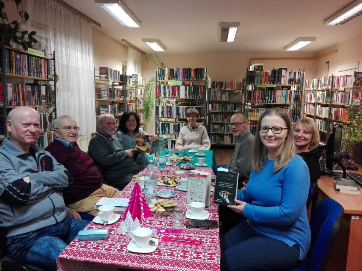 Uczestnicy DKK przy stole z książkami i słodyczami, papierowymi ozdobami; klubowicze czytający książkę, w tle zakładki DKK i książka „Kamerdyner świata” na małej sztaludze.