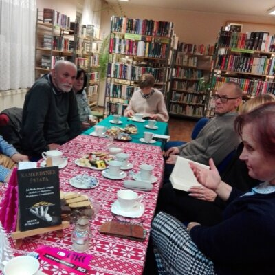 Uczestnicy DKK przy stole z książkami i słodyczami, papierowymi ozdobami; klubowicze czytający książkę, w tle zakładki DKK i książka „Kamerdyner świata” na małej sztaludze. Kliknięcie w obrazek spowoduje powiększenie.