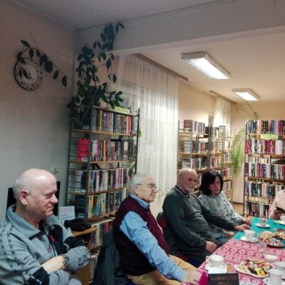 Uczestnicy DKK przy stole z książkami i słodyczami, papierowymi ozdobami; klubowicze czytający książkę, w tle zakładki DKK i książka „Kamerdyner świata” na małej sztaludze. Kliknięcie w obrazek spowoduje powiększenie.