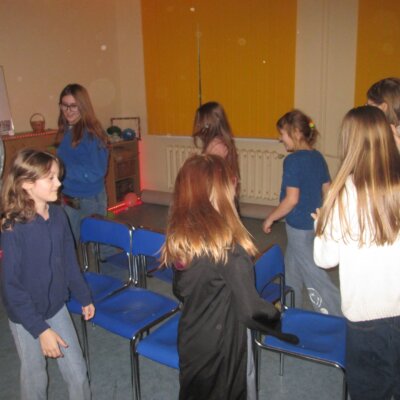 Grupa roześmianych dziewczynek tańczy wokół krzeseł na środku pomieszczenia. Klikniecie powoduje powiększenie zdjęcia.