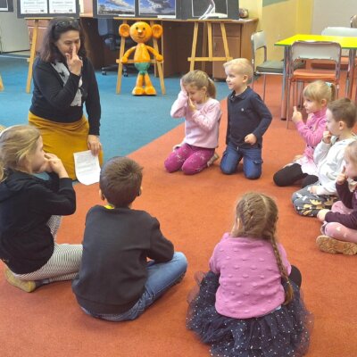 Na dywanie w kółku siedzą przedszkolaki z bibliotekarką, kobieta wskazuje palcem nos, a dzieci ją naśladują.
